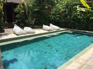 A clear swimming pool with tiled edges, surrounded by lush greenery and a tiled patio area with white cushions. - Cafe Wayan Cottages (Senggigi)