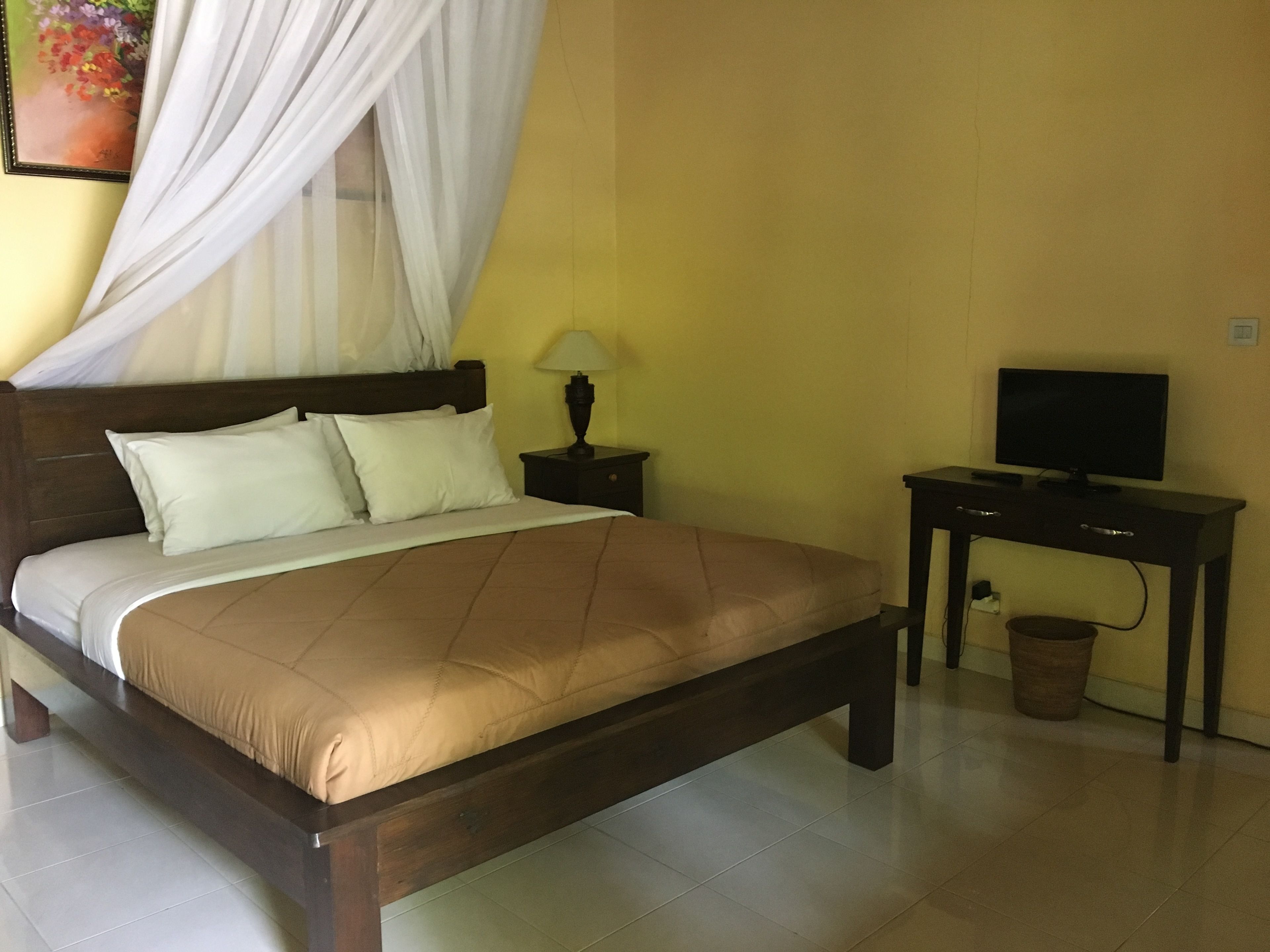 A bedroom with a bed, a bedside table with a lamp, a television on a stand, and a trash bin.