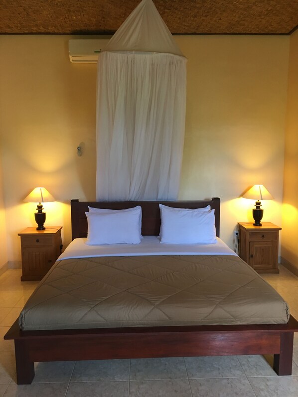 A bed with a canopy, two bedside tables with lamps, and a wall-mounted air conditioner. - Cafe Wayan Cottages (Senggigi)