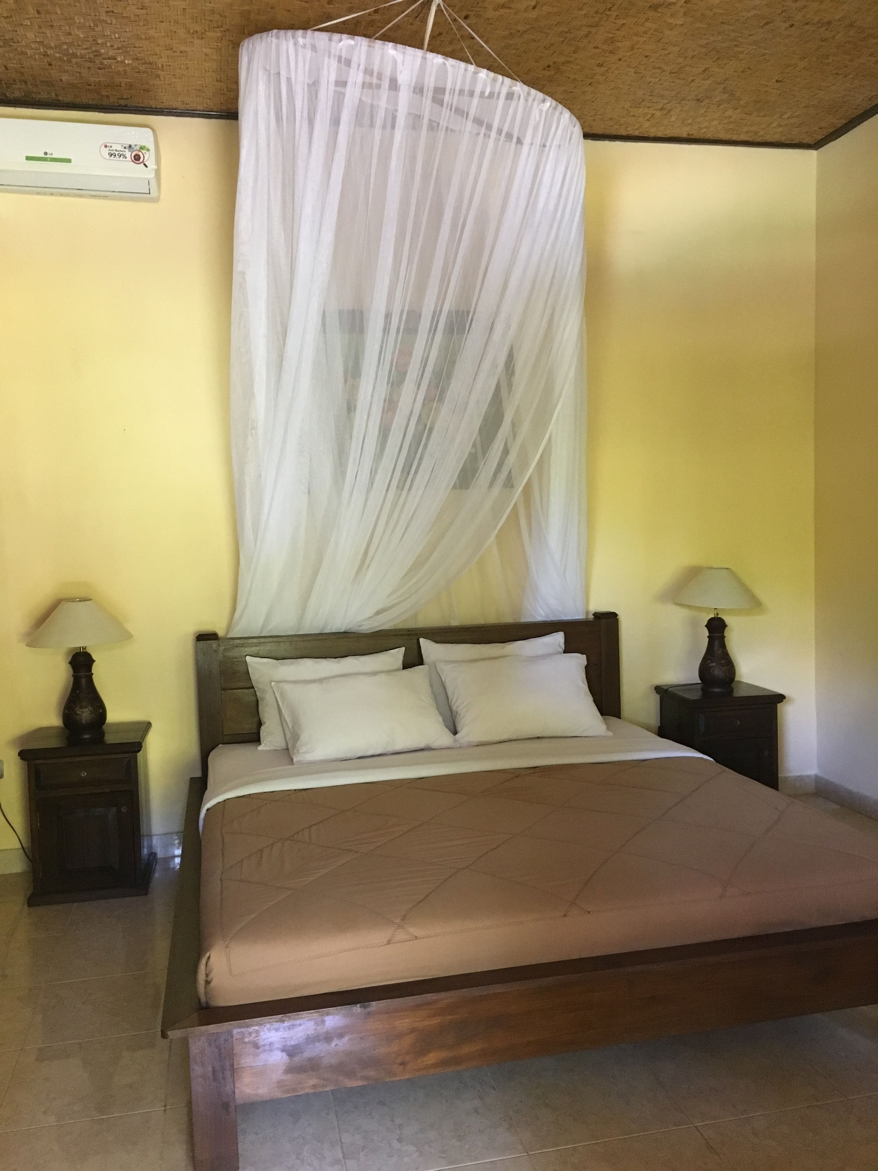 A bedroom with a bed, two bedside tables, a hanging mosquito net, and a wall-mounted air conditioner.
