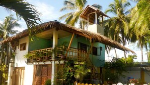 A two-story house with a thatched roof, a balcony, and a white picket fence. - Alma's Guesthouse (Panglao)