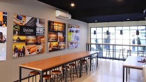 A modern indoor space with a long wooden table and multiple bar stools, large windows with a view of the outside, and walls adorned with various posters. - Livinn Hostel City Center Surabaya (Surabaya)