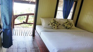 Una cama con sábanas blancas y almohadas estampadas con motivos florales, suelo de madera y vistas al verdor a través de una ventana con cortinas azules.