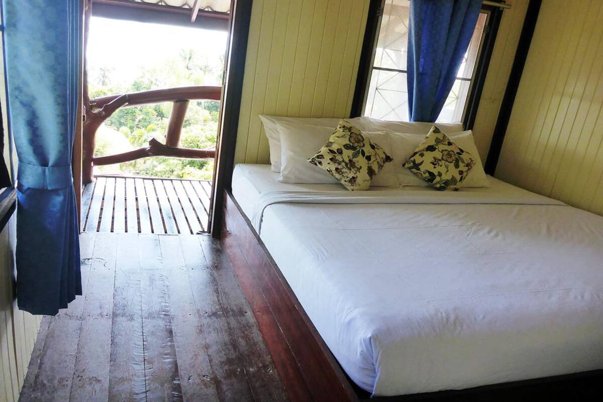 Una cama con sábanas blancas y almohadas estampadas con motivos florales, suelo de madera y vistas al verdor a través de una ventana con cortinas azules.