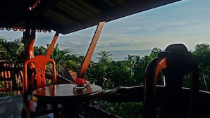Un área de estar al aire libre con mesa y sillas, con vistas a un paisaje tropical con palmeras y el océano.
