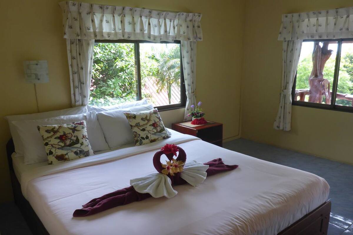 Habitación de hotel con cama, almohadones estampados, una mesita de noche y ventana con cortinas.