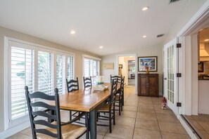 Un comedor con una mesa y sillas de madera, una ventana con persianas, y una cocina visible a través de una puerta abierta.