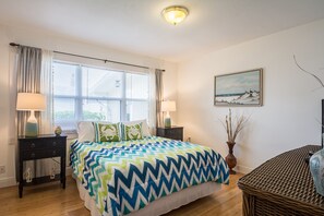 Un dormitorio con una cama, dos mesitas de noche, una lámpara, una silla de mimbre, un cuadro y un jarrón con ramas.