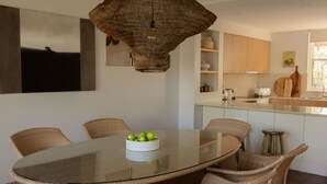 A dining area with a glass table surrounded by wicker chairs, a woven pendant light, and a bowl of green apples.