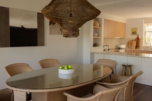 Un comedor con una mesa de vidrio rodeada de sillas de mimbre, una lámpara colgante tejida y un cuenco con manzanas verdes.
