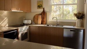 A modern kitchen with wooden cabinets, a stainless steel dishwasher, and a window with a view of trees. - Tempranillo 3br Villa within Cypress Lakes Resort (Pokolbin)