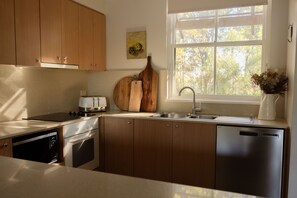 A modern kitchen with wooden cabinets, a stainless steel dishwasher, and a window with a view of trees.