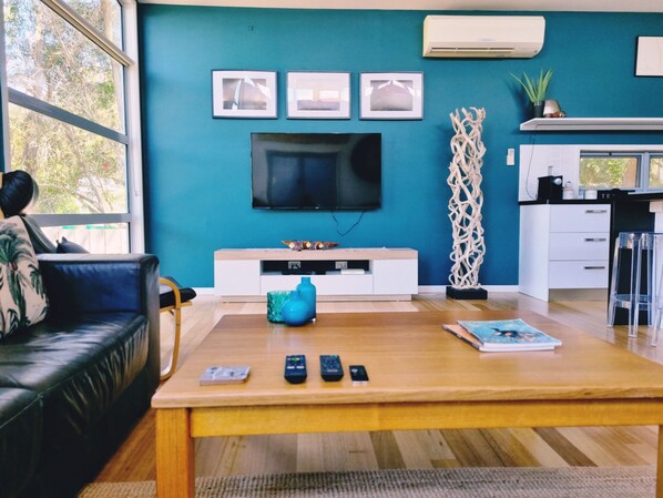 A modern living room with a wooden coffee table, a black leather sofa, a flat-screen TV mounted on a teal wall, and a white kitchen area with a countertop and cabinets.