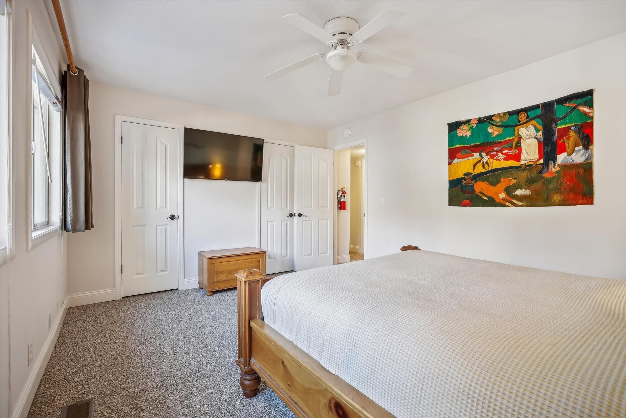 A bedroom with a bed, a ceiling fan, a painting on the wall, and a door leading to another room.