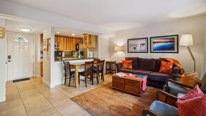 A living room with a sofa, coffee table, and chairs, a kitchen with wooden cabinets and appliances, and two framed pictures on the wall.