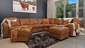 A modern living room with a brown leather sectional sofa, a large framed picture of a cow, and a view of a garden through a sliding glass door. - The Hidden Door Bowral Villa - the Heart of Bowral (Bowral)