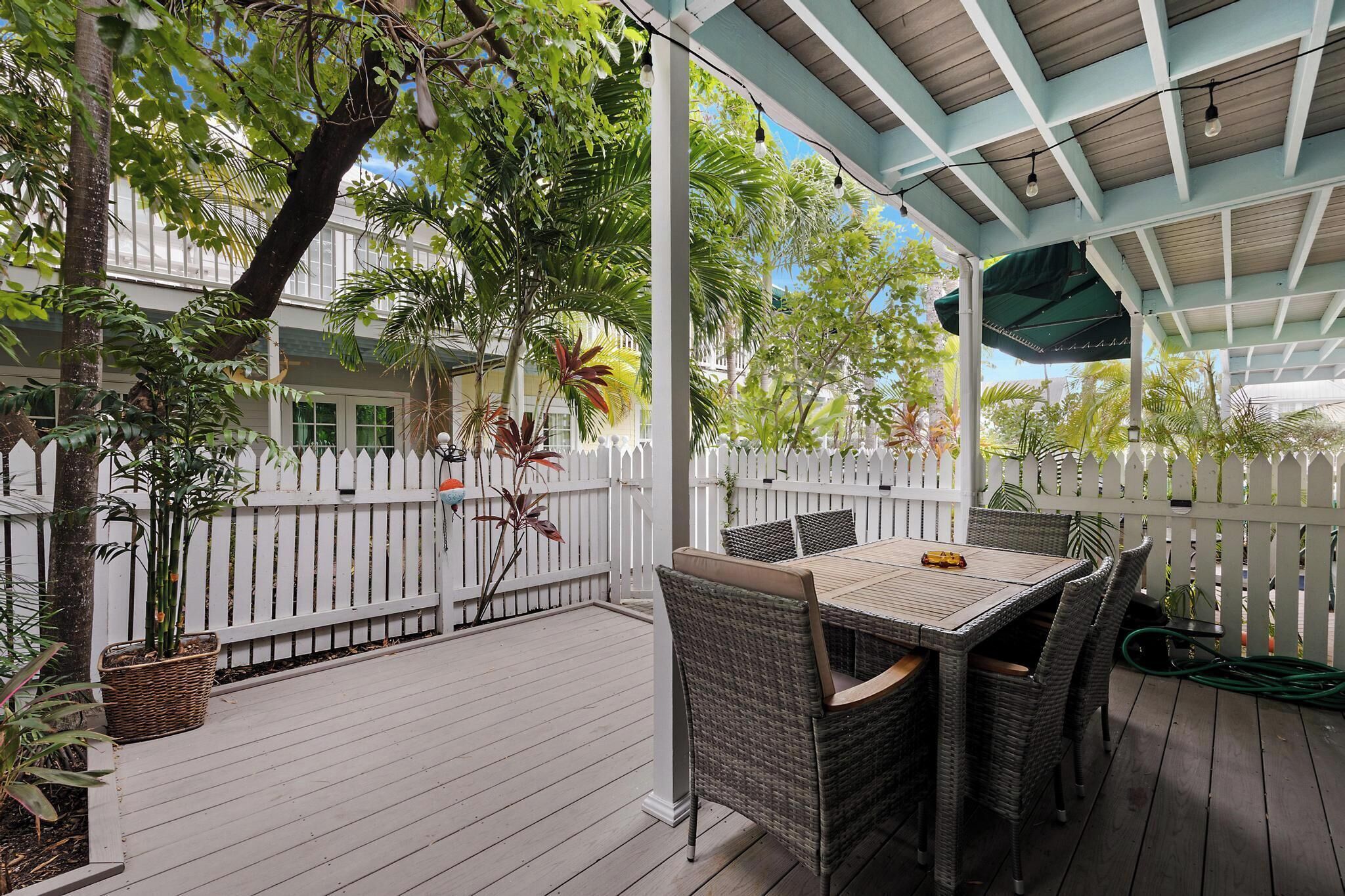 Eine überdachte Terrasse im Freien mit Korbmöbeln, einem weißen Zaun und üppiger Bepflanzung.