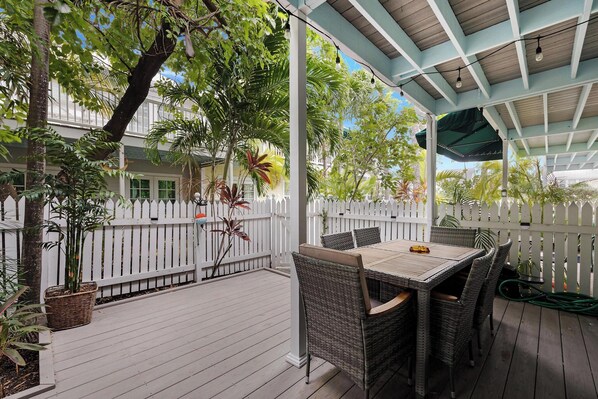 A covered outdoor patio with wicker furniture, a white picket fence, and lush greenery.