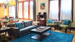A spacious living room with a leather sofa, a coffee table, and two armchairs. There is a dining area with a table and chairs, and a bookshelf with speakers.