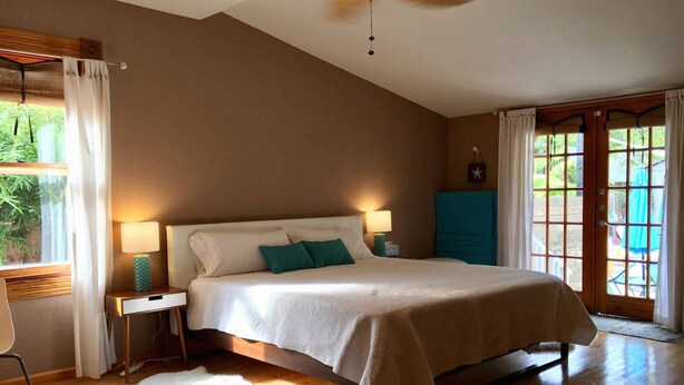 A bedroom with a large bed, two bedside lamps, a wooden floor, a white furry rug, and a view of greenery outside.