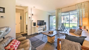 Una sala de estar moderna con un sofá, un sillón y una mesa de centro. Hay un televisor sobre un mueble blanco y un ventanal con cortinas.