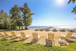A beachside seating area with lounge chairs and picnic tables, a wooden pier extending into the water, and a clear view of the sea and distant mountains. - Panoramic Lake Views & Modern Comfort - Spacious Rocky Ridge #37 (Tahoe City)