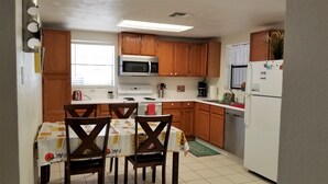 Una pequeña cocina con armarios de madera, un microondas, una estufa y un refrigerador. Hay una mesa de comedor con sillas y una ventana con persianas.