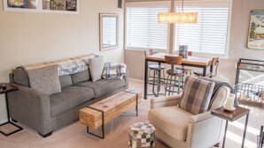 Ein gemütliches Wohnzimmer mit grauem Sofa, einem Holztisch, einem weißen Sessel und einem Essbereich mit Holztisch und Barhocker.