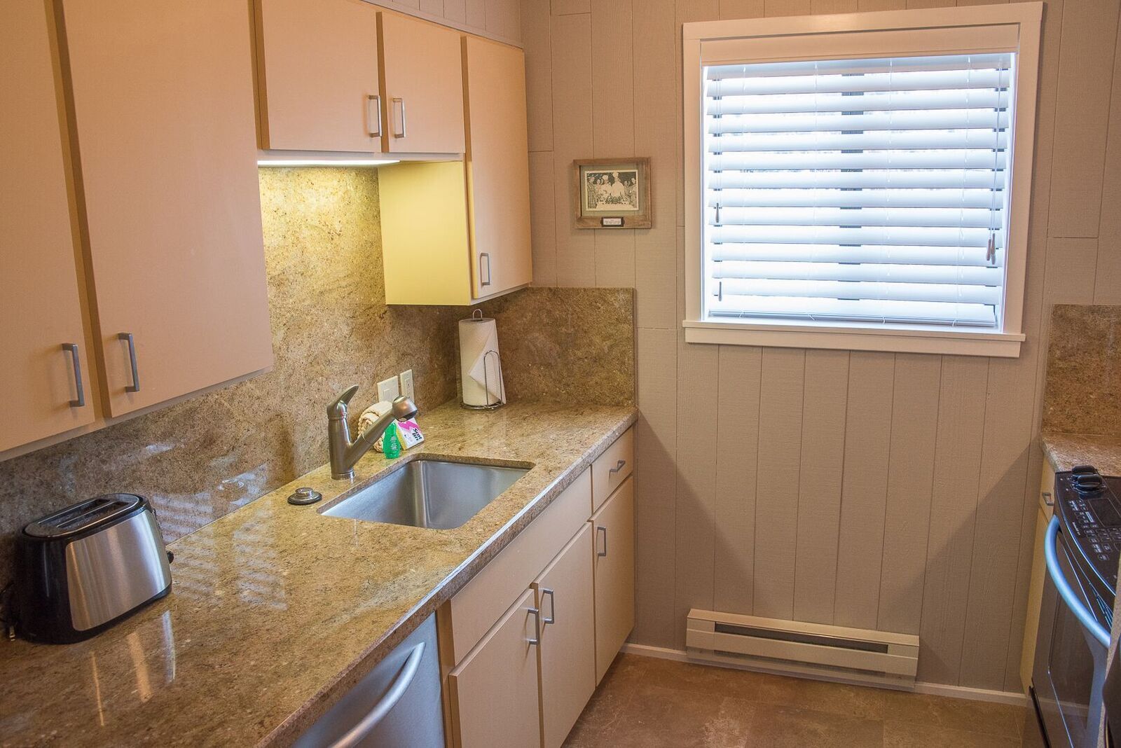 Una cocina con encimeras de granito, un fregadero de acero inoxidable, un tostador y una ventana con persianas.