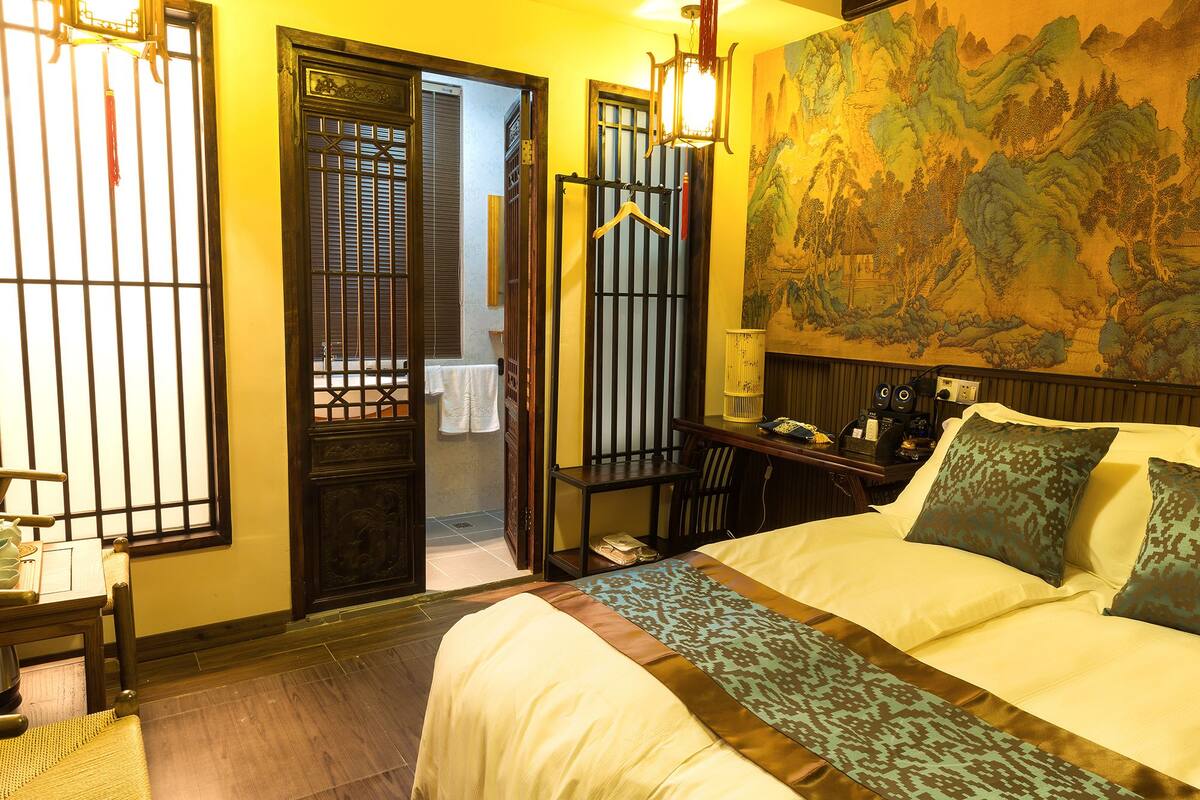 Una habitación de hotel con cama, escritorio con lámpara, mapa en la pared y puerta tradicional con ventanal.