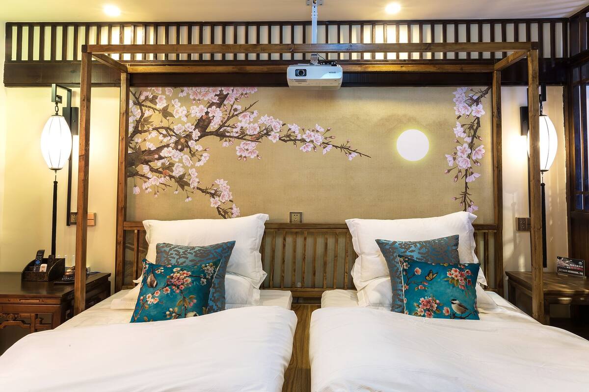 Habitación de hotel con una cama de estilo japonés tradicional, dos almohadas con motivos florales, un proyector montado en la pared y una estructura de techo de madera.