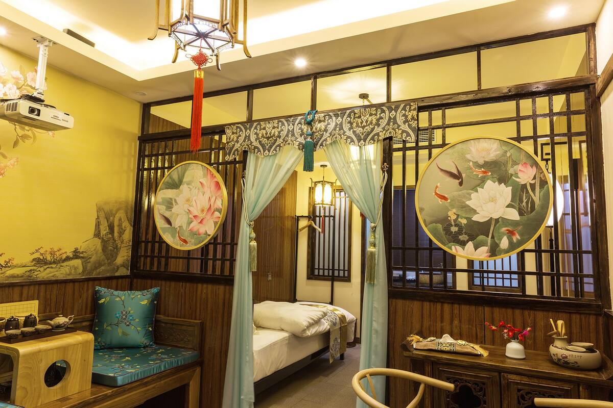 Una habitación tradicional con cama, un banco de madera, una pequeña mesa con vajilla para té y un biombo decorativo con motivos florales.