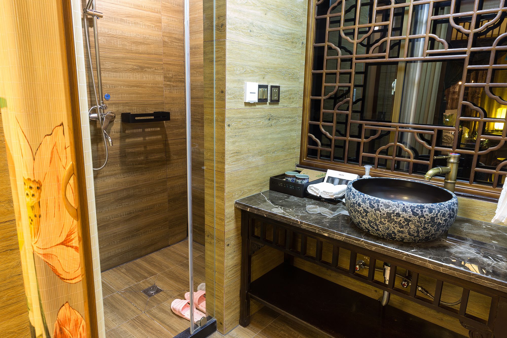A bathroom with a wooden floor, a glass shower enclosure, a sink with a decorative bowl, and a mirror with a traditional lattice design.