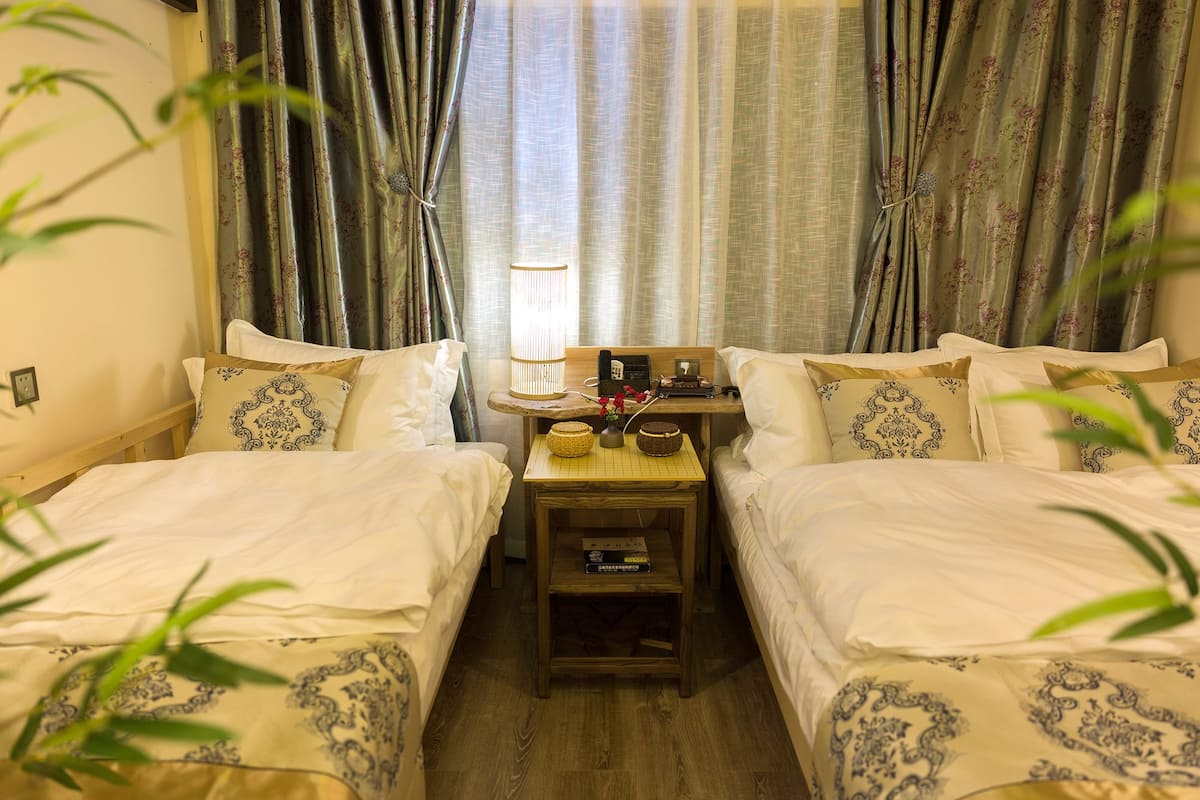 Una habitación de hotel con dos camas, una mesita de madera, una lámpara y una ventana con cortinas.