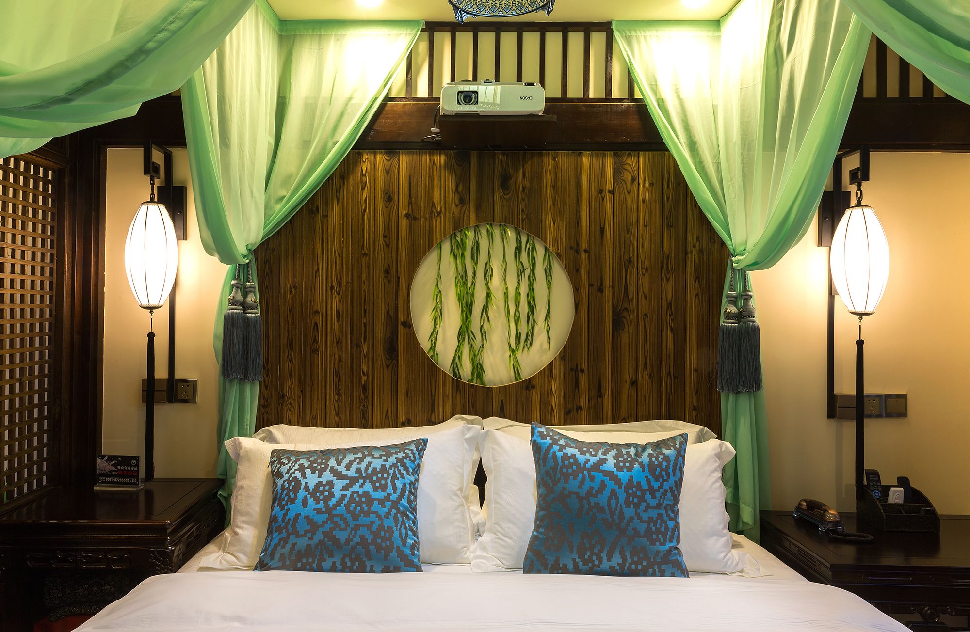 Een hotelkamer met een houten hoofdeinde, een projecter en een bed met kussens in een wit en blauw patroon.