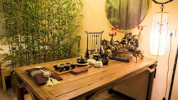 A wooden table with various tea sets, a decorative Buddha, and a bonsai tree in a room with a bamboo wall and a round mirror.