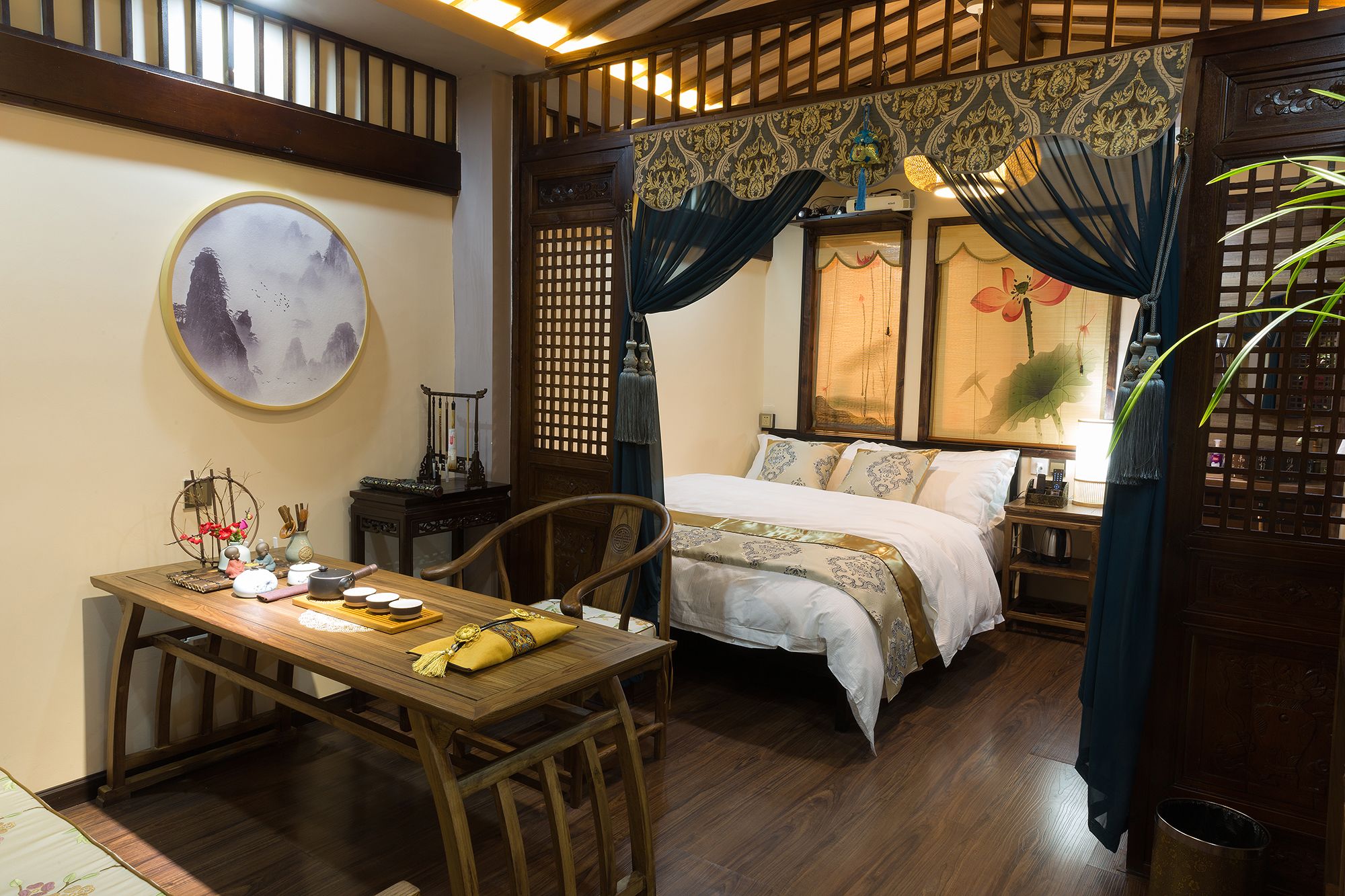 A traditional room with a bed, a dining table, and a chair. There is a painting on the wall and a window with curtains.