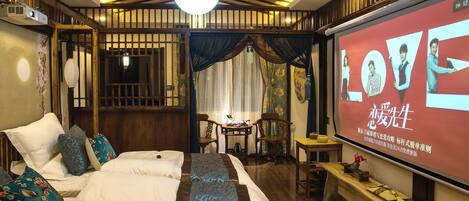 A traditional-style bedroom with a bed, pillows, a desk, and a screen displaying a movie.