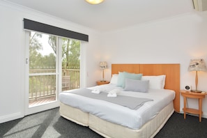 A bedroom with a large bed, two bedside tables with lamps, a window with a view of trees, and a balcony.