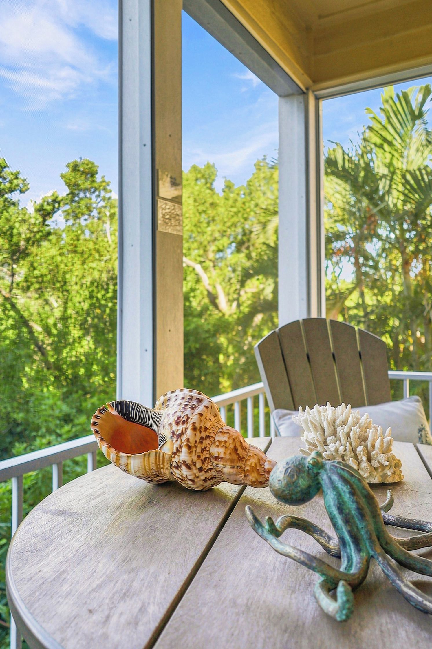 Ein Holztisch mit Muscheln und einem Kaktus-Ornament, ein Stuhl und der Blick auf Bäume durch ein Fenster.