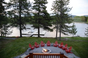 Una terraza junto al lago con sillas Adirondack rojas dispuestas alrededor de un pozo de fuego, con vistas a un lago tranquilo con muelle y barcos.