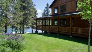 A wooden cabin with a covered deck overlooking a lake, surrounded by trees and grass.