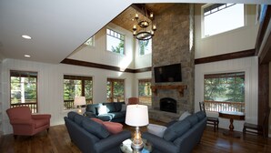 A spacious living room with a stone fireplace, large windows, and a high ceiling.