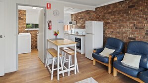 Cocina compacta con pared de ladrillo, un refrigerador blanco y un microondas. Hay una mesa de comedor de madera con dos taburetes y una silla de cuero azul.