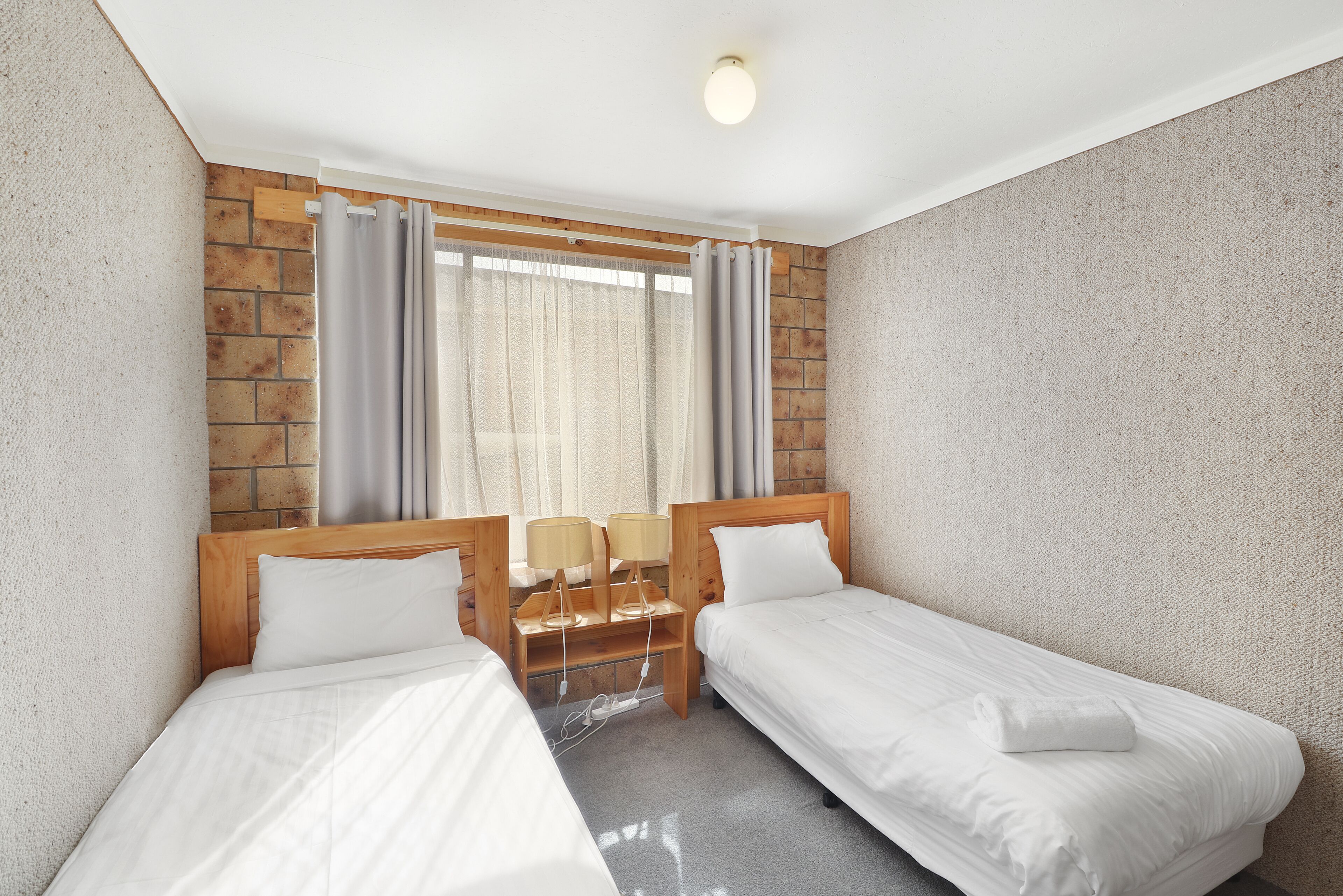 Habitación de hotel con dos camas individuales, cabecera de madera, mesita de noche con lámparas y ventana con cortinas.