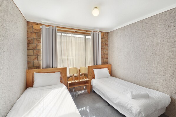 Une chambre d’hôtel avec deux lits simples, une tête de lit en bois, une table de chevet avec des lampes et une fenêtre avec des rideaux.