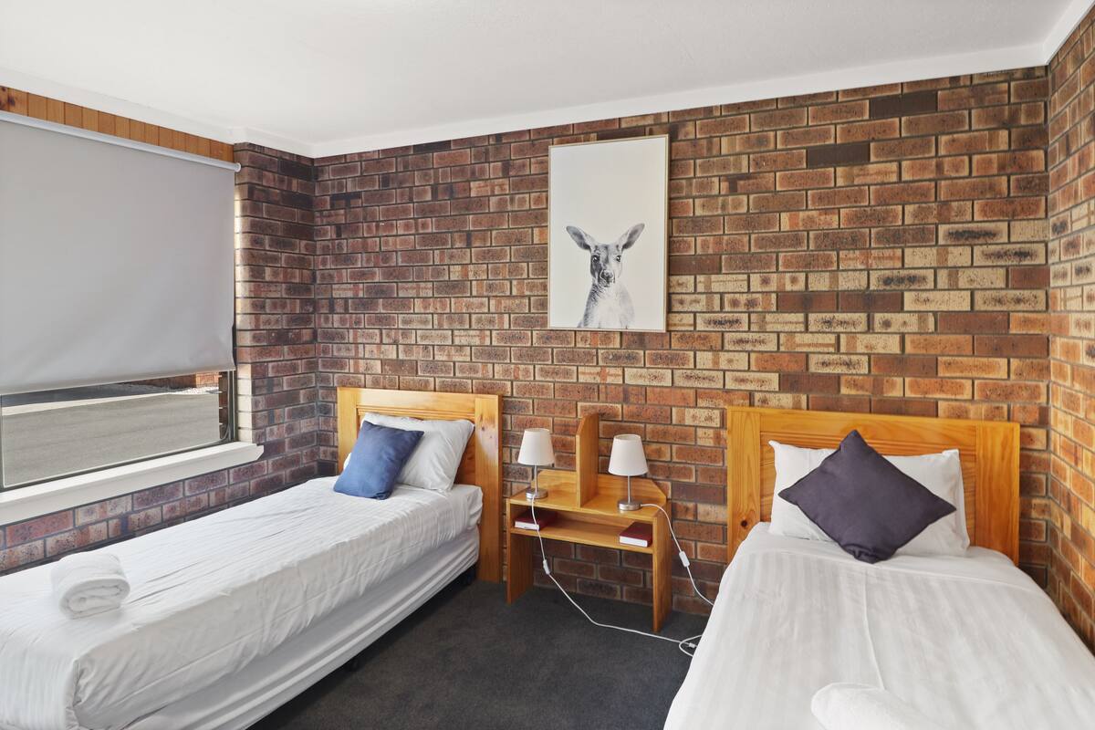 A hotel room with two single beds, a brick-patterned wall, a window with a blind, and a framed picture above the beds.