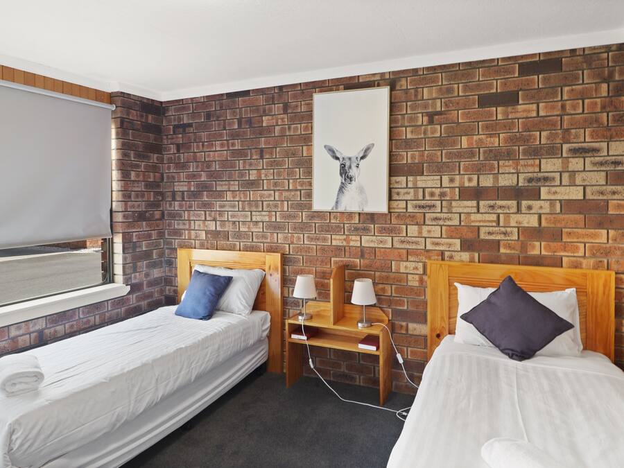 A hotel room with two single beds, a brick-patterned wall, a window with a blind, and a framed picture above the beds.