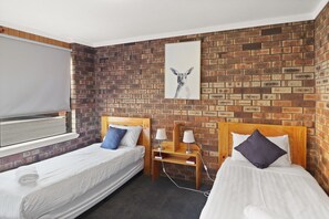 A hotel room with two single beds, a brick-patterned wall, a window with a blind, and a framed picture above the beds. - Kellraine Holiday Units (St Helens)