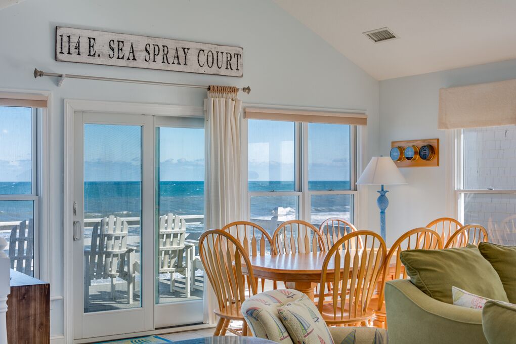 A room with a view of the ocean, a dining table with chairs, and a sign reading '114 E. SEA SPRAY COURT.'